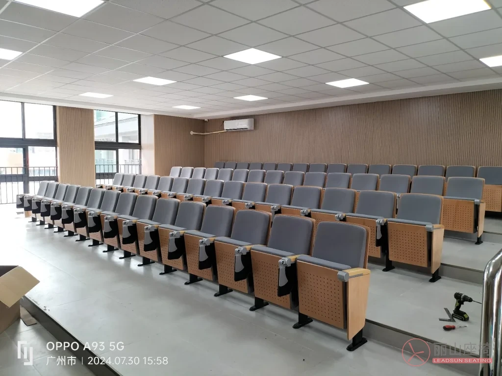 罗边小学礼堂全景实景图，灰色软包座椅配浅木色背板，阶梯式排列，大面积自然采光